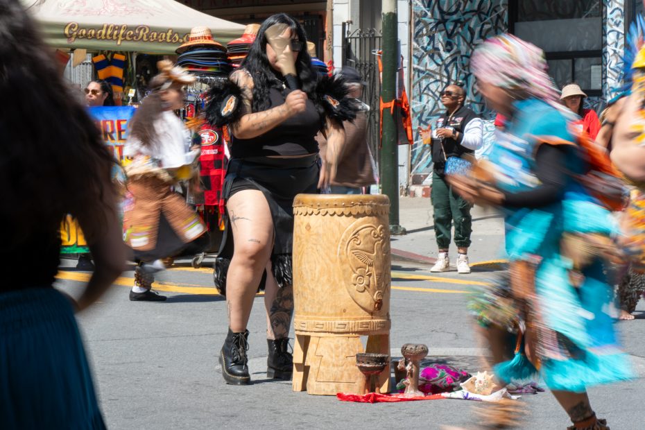 A person in a feathered costume plays a large wooden drum on a street. People in colorful outfits dance around them, creating a lively atmosphere.
