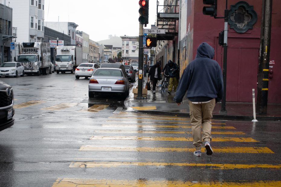Person in a hoodie crosses a wet street at a yellow-striped crosswalk on a cloudy day in an urban area.