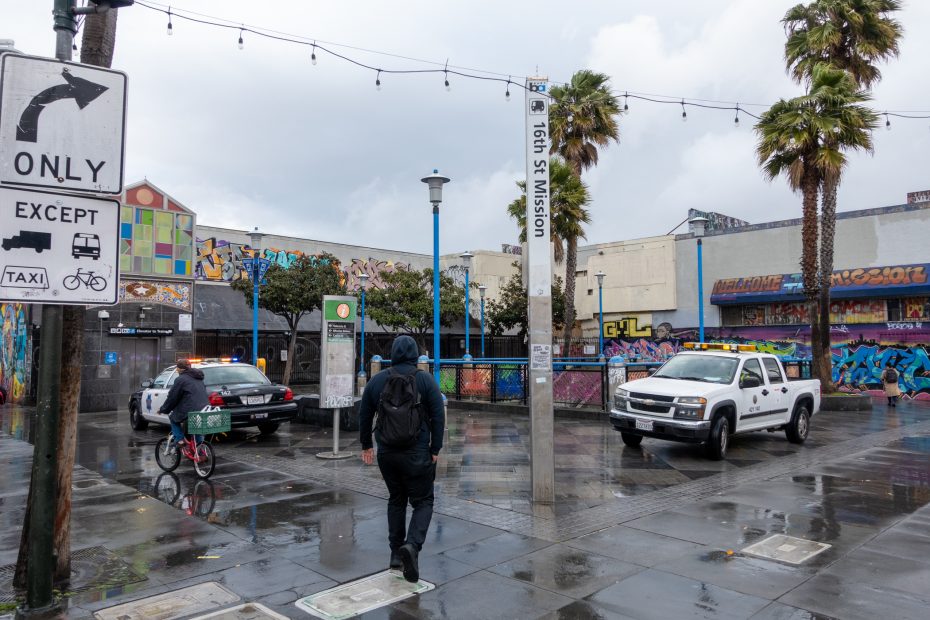 A person walks by police and utility vehicles near the 16th St Mission BART station on a rainy day. Grafitti covers nearby walls. Palm trees and streetlights line the sidewalk.
