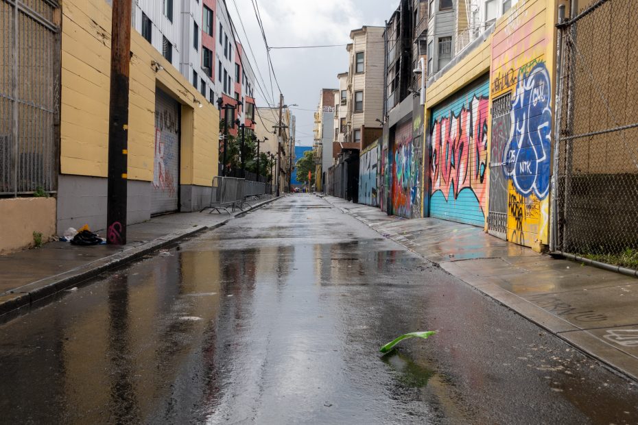Narrow urban alley with wet pavement, colorful graffiti on buildings, and overhead power lines. A single green leaf lies on the ground.