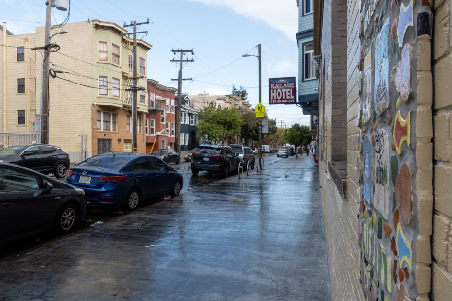 Wet urban street with parked cars, multi-story buildings, and utility poles. A hotel sign is visible on the right. Sidewalk features colorful tile artwork. Overcast sky above.