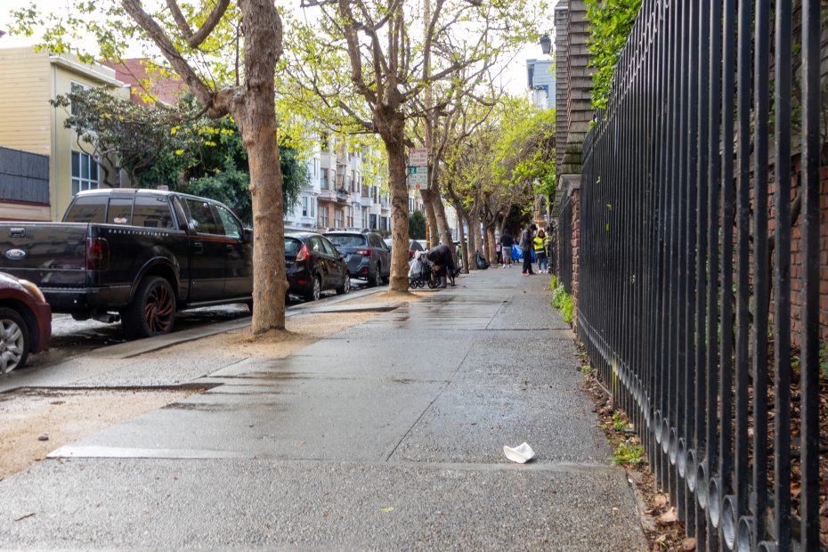 Wet city sidewalk lined with trees and parked cars; a person with a stroller and another person in yellow vest walk in the distance. Black metal fence runs along the right side.
