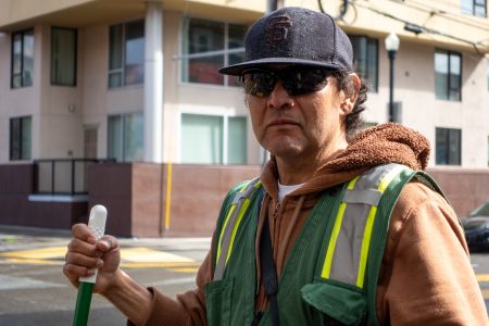 Person wearing a hoodie, cap, and sunglasses holds a broom on a city street, standing in front of a building.
