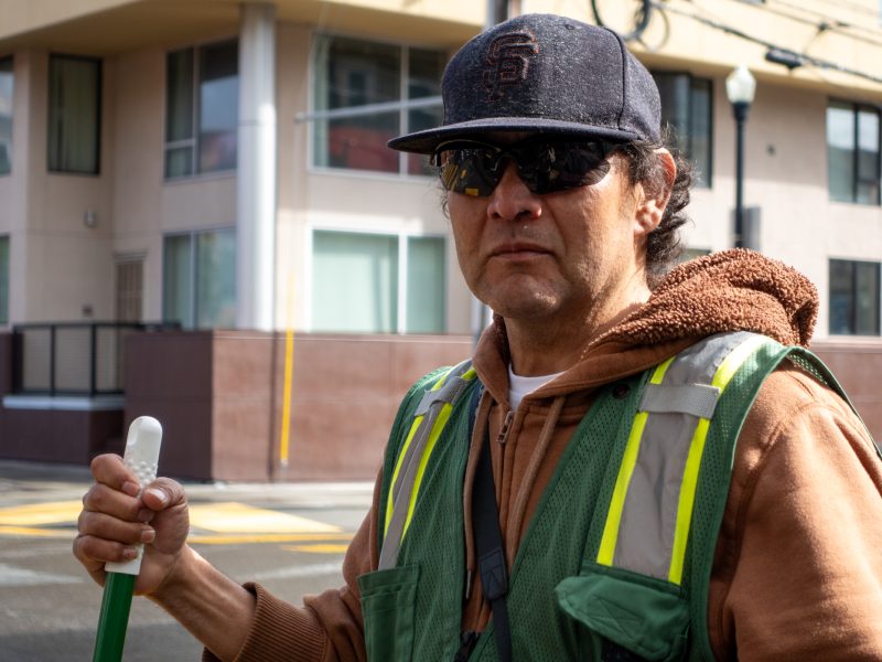Person wearing a hoodie, cap, and sunglasses holds a broom on a city street, standing in front of a building.