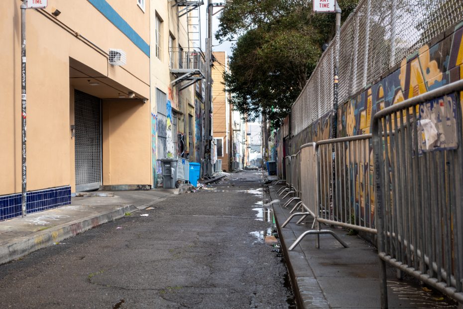 Narrow urban alley with graffiti, puddles, and chain-link fence, flanked by buildings and metal barricades.