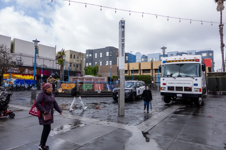A street scene on a rainy day in a plaza with people walking, a Mobile Command Two truck parked, and colorful murals on surrounding buildings.
