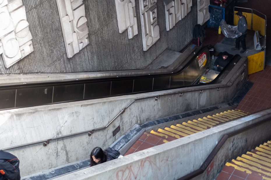 A person descends an indoor escalator, while another person at the bottom appears to be heading up. The area is industrial with concrete walls and a few people nearby.