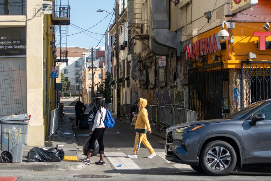 People walking on a city street near a taqueria, with a parked car and buildings in the background.
