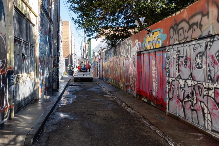 A truck parked in a graffiti-covered alley with a person standing nearby. Walls on both sides are heavily painted with colorful graffiti.