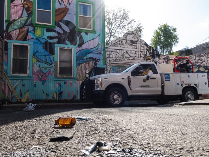 Utility truck parked by a colorful graffiti wall, with debris including a brown bottle on the ground, under a partly cloudy sky.