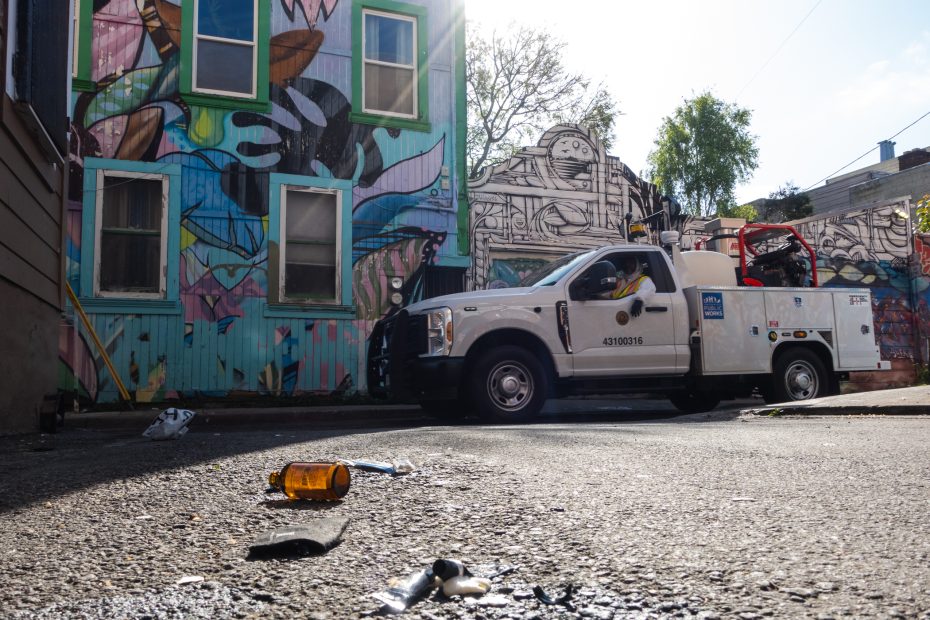Utility truck parked by a colorful graffiti wall, with debris including a brown bottle on the ground, under a partly cloudy sky.