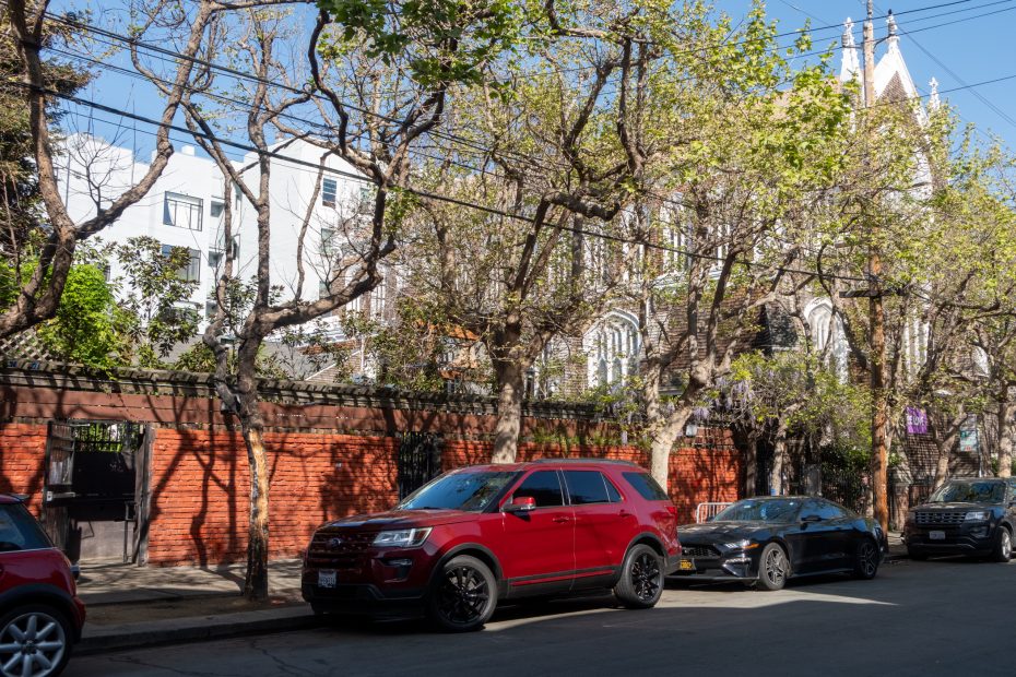 Street with parked cars under trees, including a red SUV and a black car, in front of a brick wall and a partially visible church building.