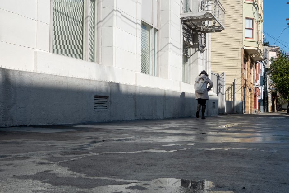 Person in a gray jacket walking down an urban street with wet pavement and a few puddles, near a white building with fire escape stairs.