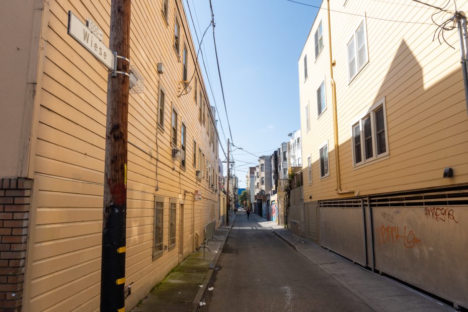Narrow urban alley with beige buildings on both sides, utility wires crossing above, and graffiti on some walls under a clear blue sky.