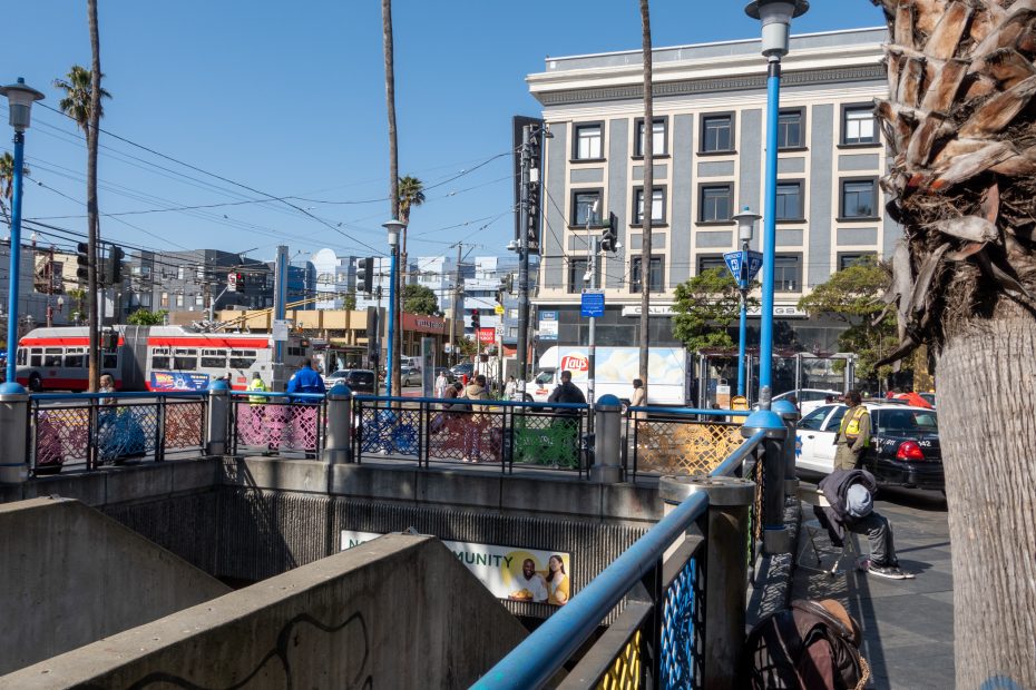 Urban street scene with people walking near a subway entrance, surrounded by buildings and palm trees. A police car and a trolley are visible in the background.