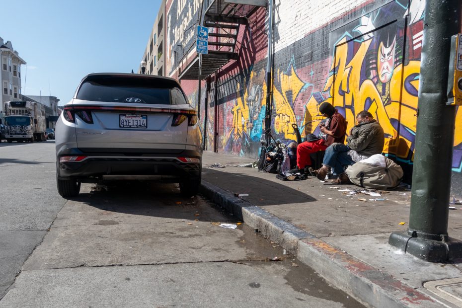 A silver car parked on a city street near a graffiti-covered wall. Two people sit on the sidewalk surrounded by belongings. A street sign and tall buildings are in the background.