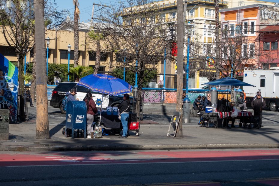 Street vendors with blue umbrellas sell items near a mailbox on a sunny day, with colorful buildings and trees in the background.