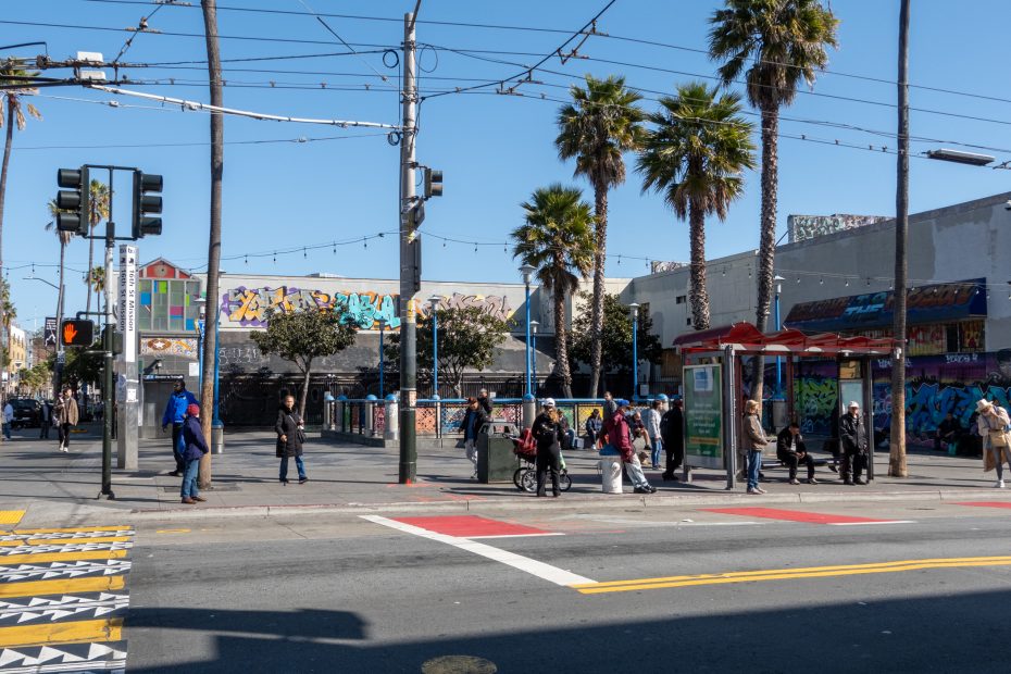 A street scene with pedestrians, palm trees, and a wall with colorful graffiti. People stand near a bus stop while cars wait at a crosswalk. Overhead wires stretch across the blue sky.