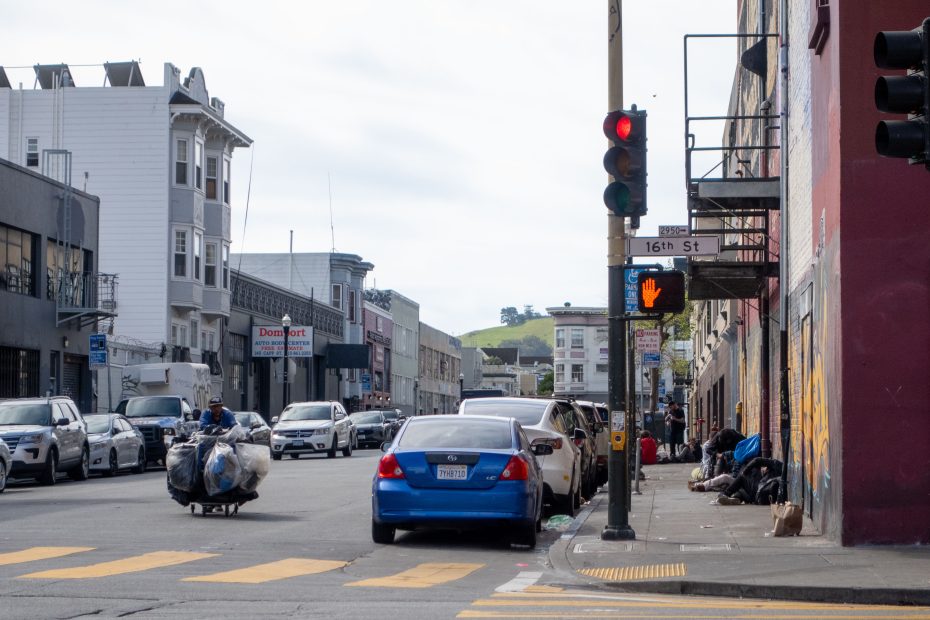 Urban street view with buildings lining both sides, parked cars, a blue car driving, a person pushing a cart with bags, pedestrians on the sidewalk, and a red traffic light at the intersection.