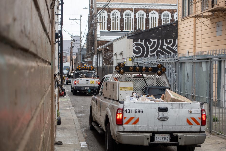 Three utility trucks are parked in a narrow urban alleyway with trash and equipment in their beds, surrounded by buildings and power lines.