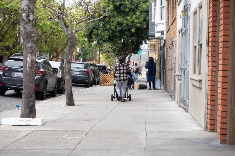 Person pushing a stroller down a city sidewalk lined with parked cars and trees; other people are visible further down the sidewalk near buildings.