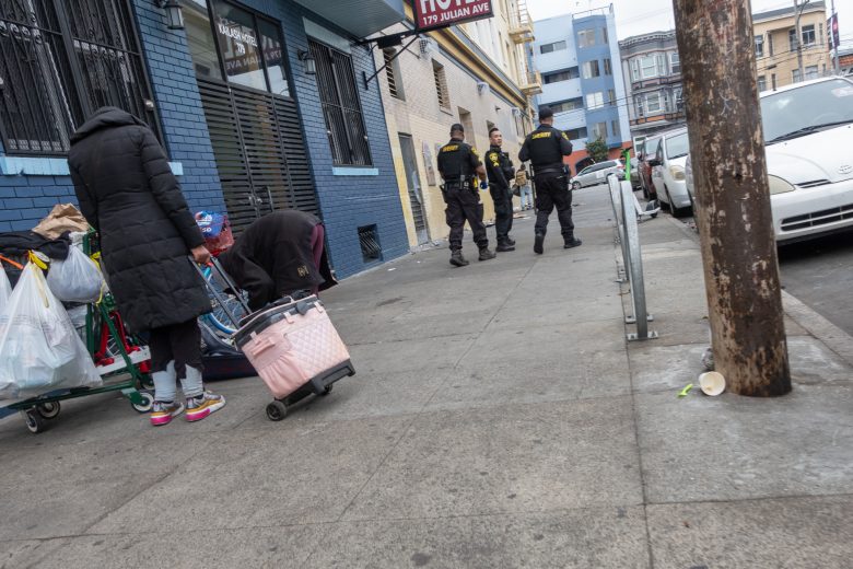 Two people with carts stand on a city sidewalk, one bending over a pink cart, while four uniformed officers stand further away near a building.