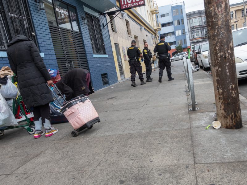 Two people with carts stand on a city sidewalk, one bending over a pink cart, while four uniformed officers stand further away near a building.