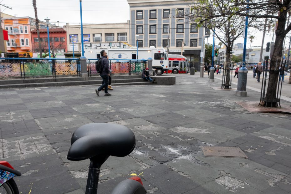 A city plaza with a bicycle seat in the foreground, a few people walking or sitting, and a white bus parked near colorful buildings in the background.