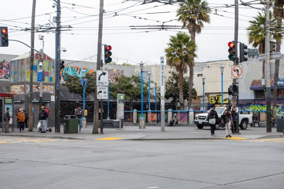Street intersection with people waiting near a public transit stop; graffiti covers surrounding buildings, and palm trees line the sidewalk. Traffic lights and street signs are visible.