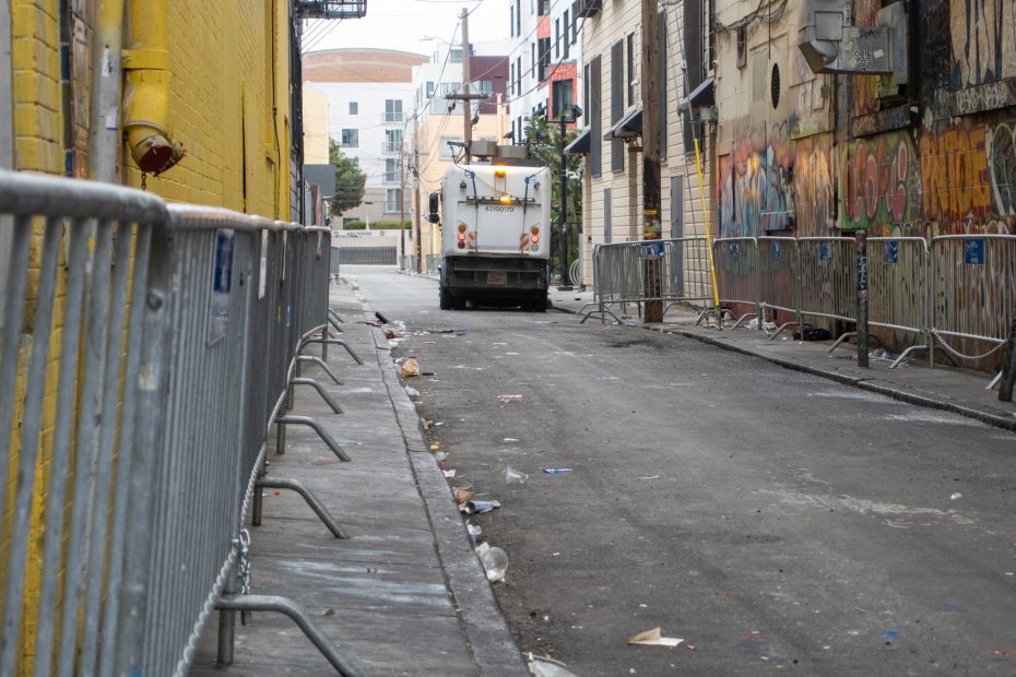 A street sweeper cleans a trash-littered alley lined with metal barricades and graffiti-covered walls.