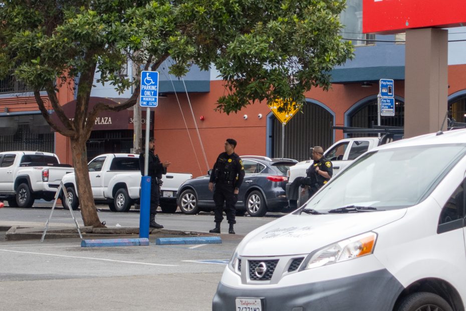 Two uniformed police officers stand in a parking lot near a tree and several parked vehicles, including a white van with a California license plate.