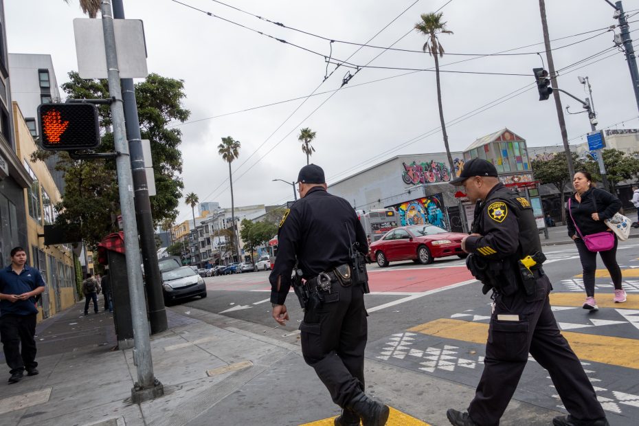 Two police officers cross a city street at a crosswalk under a red hand signal, with pedestrians and graffiti-covered buildings in the background.