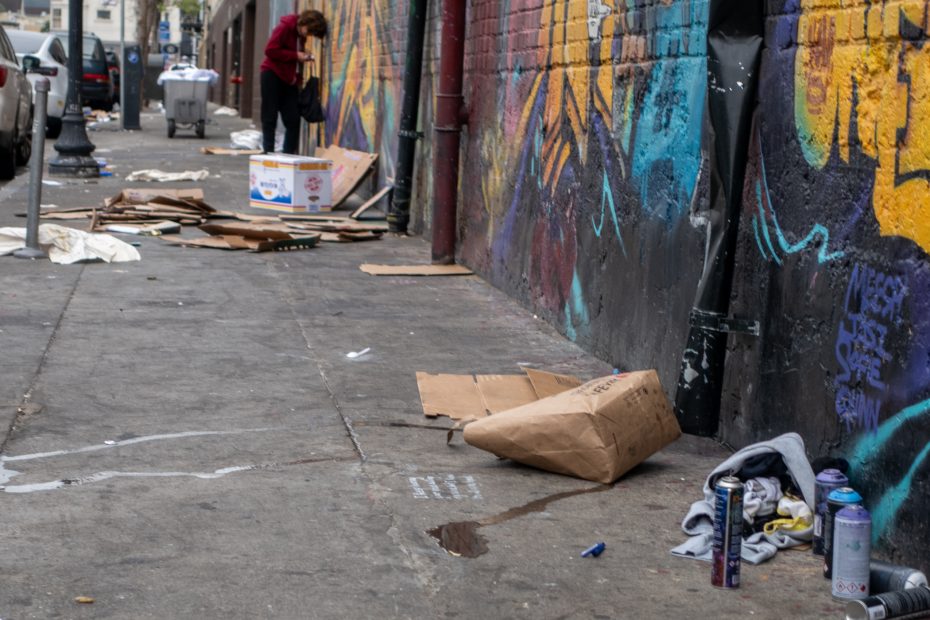 A person sorts through cardboard boxes on a littered urban sidewalk beside a graffiti-covered wall, with cans and trash scattered around.