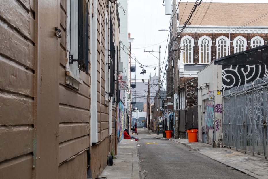 A narrow urban alleyway with graffiti-covered walls, utility wires overhead, orange trash bins, and a few people sitting along the sides.