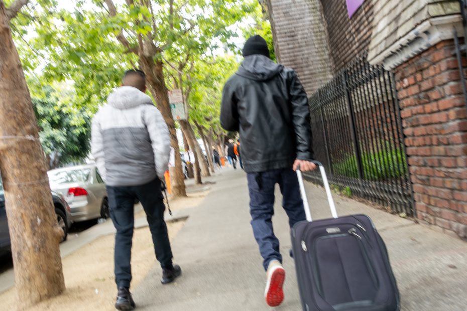 Two people walk down a tree-lined sidewalk; one pulls a wheeled suitcase and both wear jackets. Cars are parked along the street.
