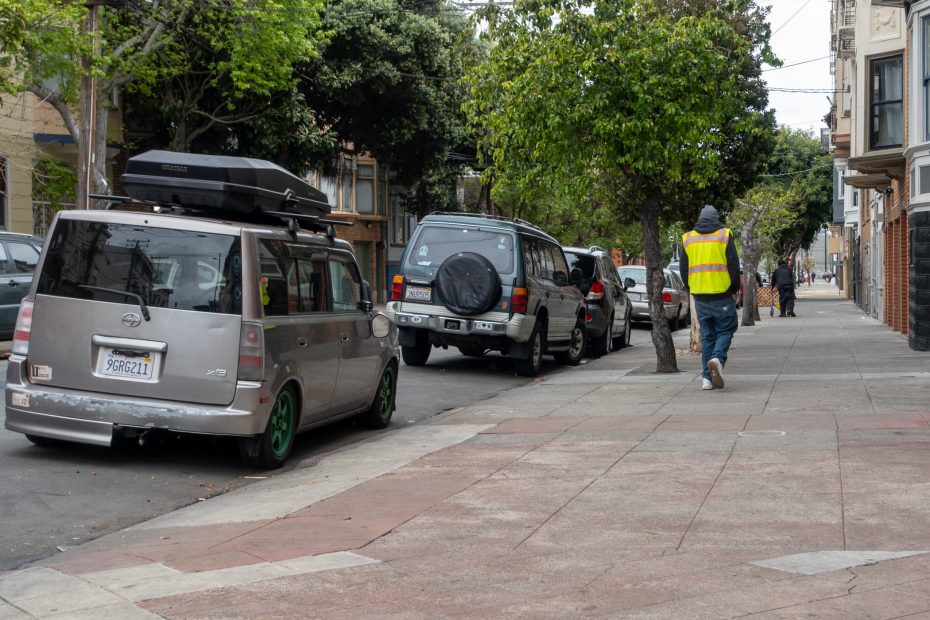 A person wearing a yellow safety vest walks on a city sidewalk next to a row of parked cars and trees.