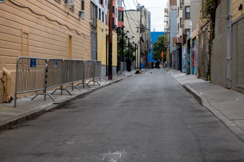 A narrow urban alleyway lined with painted buildings and metal barricades, with scattered debris visible on the street and distant figures near the end.