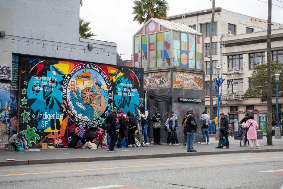 A group of people stand and sit near a colorful mural reading "American Indian Cultural District" on a city street with a painted glass structure in the background.