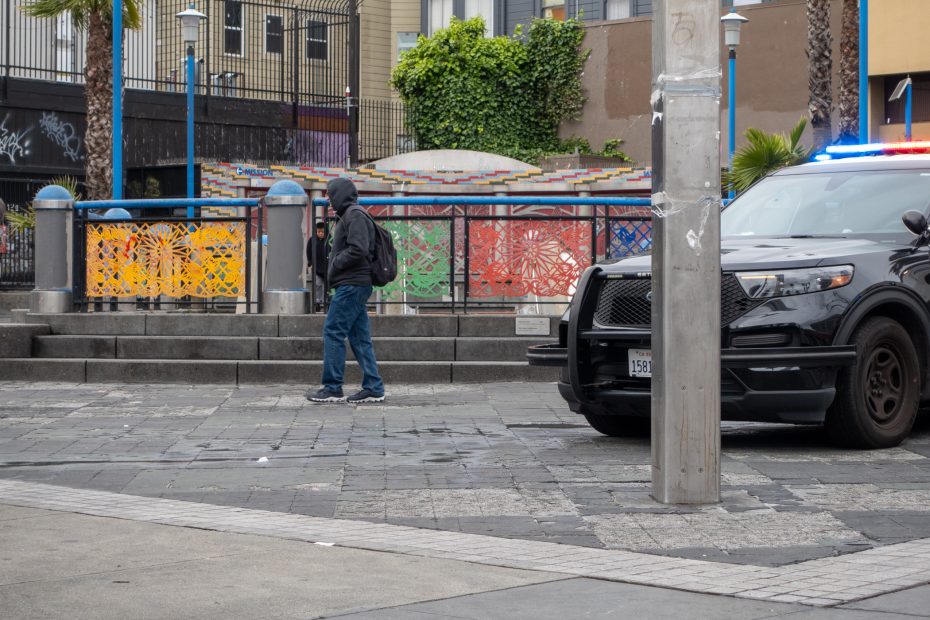 A person with a backpack walks past a black police SUV with flashing lights near colorful tiled steps in an urban area.