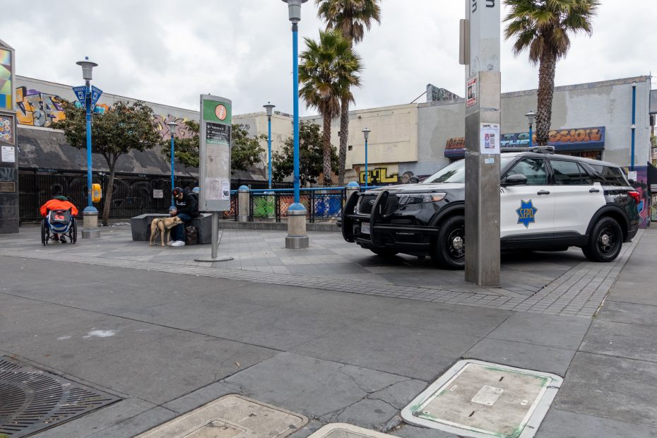 A police SUV is parked in an urban plaza near people sitting on benches and a stroller; graffiti art is visible on surrounding buildings.