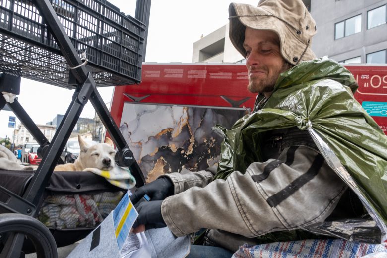 A man wearing a hooded jacket sits outdoors near a cart with a small dog lying on a blanket, and various personal belongings are visible around them.