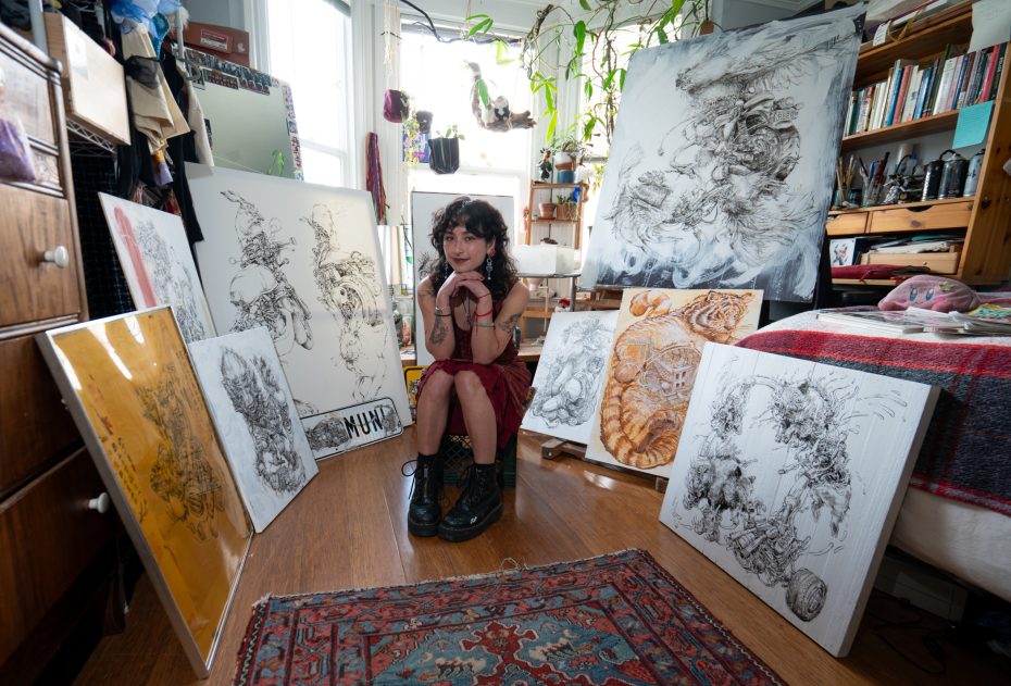 Artist seated on the floor surrounded by intricate artwork on canvas in a cluttered, plant-filled studio space with shelves and a carpet.