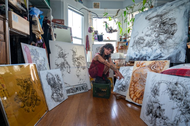 Artist sitting on a crate in a studio, surrounded by large canvases featuring detailed animal artwork. Sunlight streams through a window, and plants hang from the ceiling.