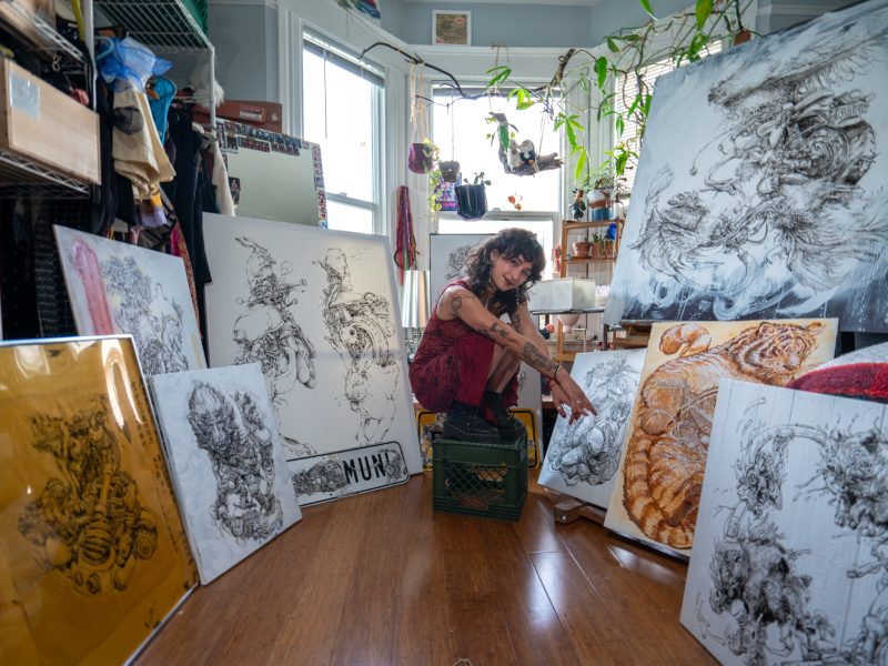 Artist sitting on a crate in a studio, surrounded by large canvases featuring detailed animal artwork. Sunlight streams through a window, and plants hang from the ceiling.