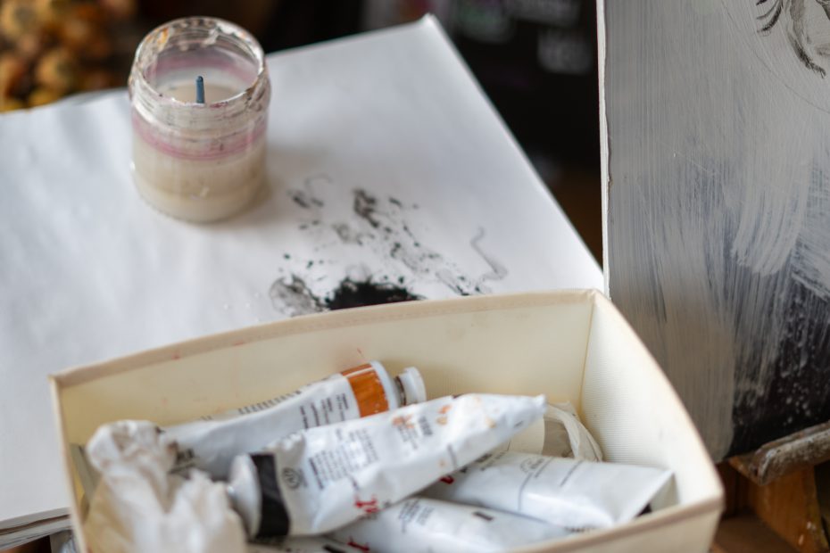 Art supplies including paint tubes, a canvas with black paint strokes, and an open jar are arranged on a table.