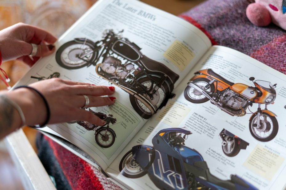 Hands with rings flipping through a motorcycle catalogue, featuring detailed images and descriptions of various bikes on a textured surface.