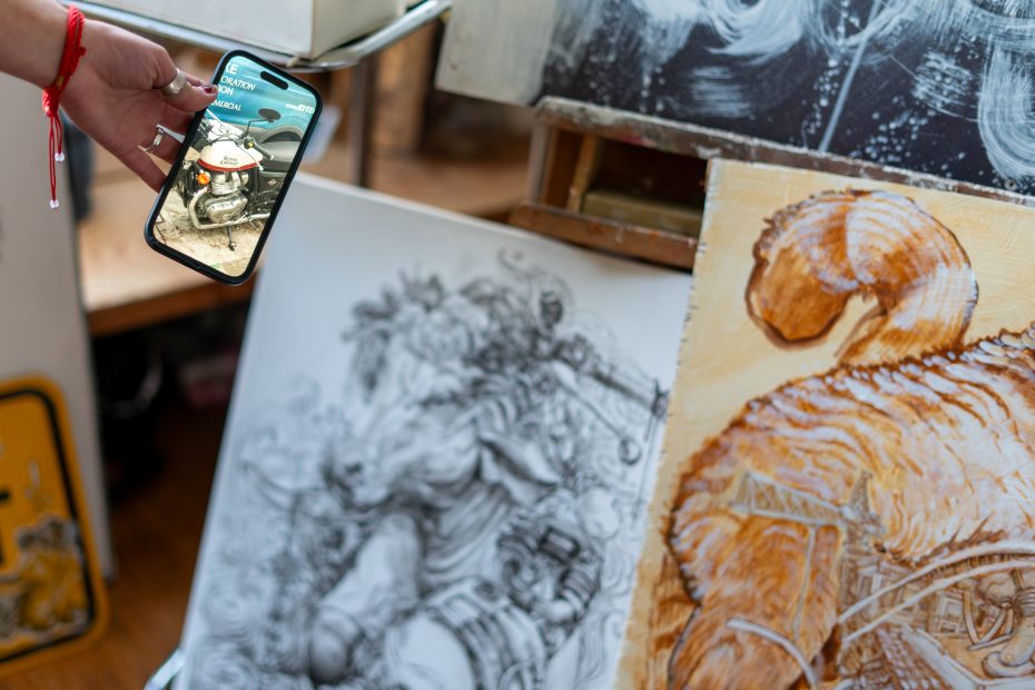 A person holds a smartphone displaying an image of a motorcycle, in front of detailed artwork featuring sketches and paintings, including a figure and an animal in orange hues.