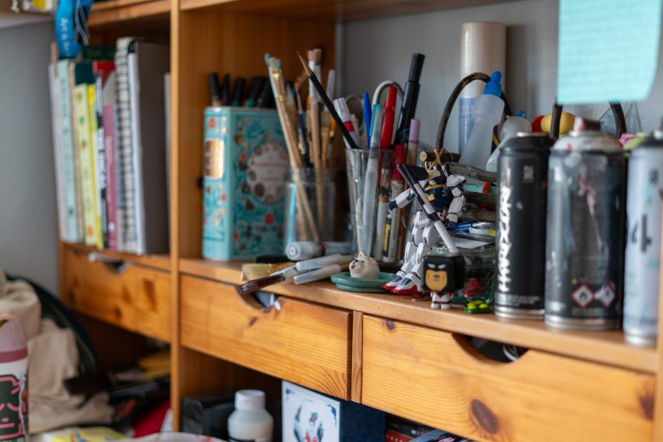 Wooden shelf cluttered with art supplies, books, cans, paintbrushes in jars, a small robot figurine, and various stationery items.