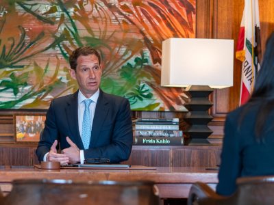 A person in a suit talks at a desk with books and a lamp in the background.