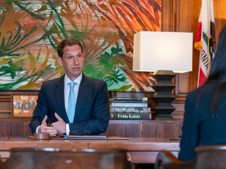 A person in a suit talks at a desk with books and a lamp in the background.